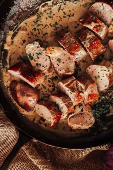 Sliced pork tenderloin in a creamy sauce with herbs in a pan on a wooden table with a burlap cloth.