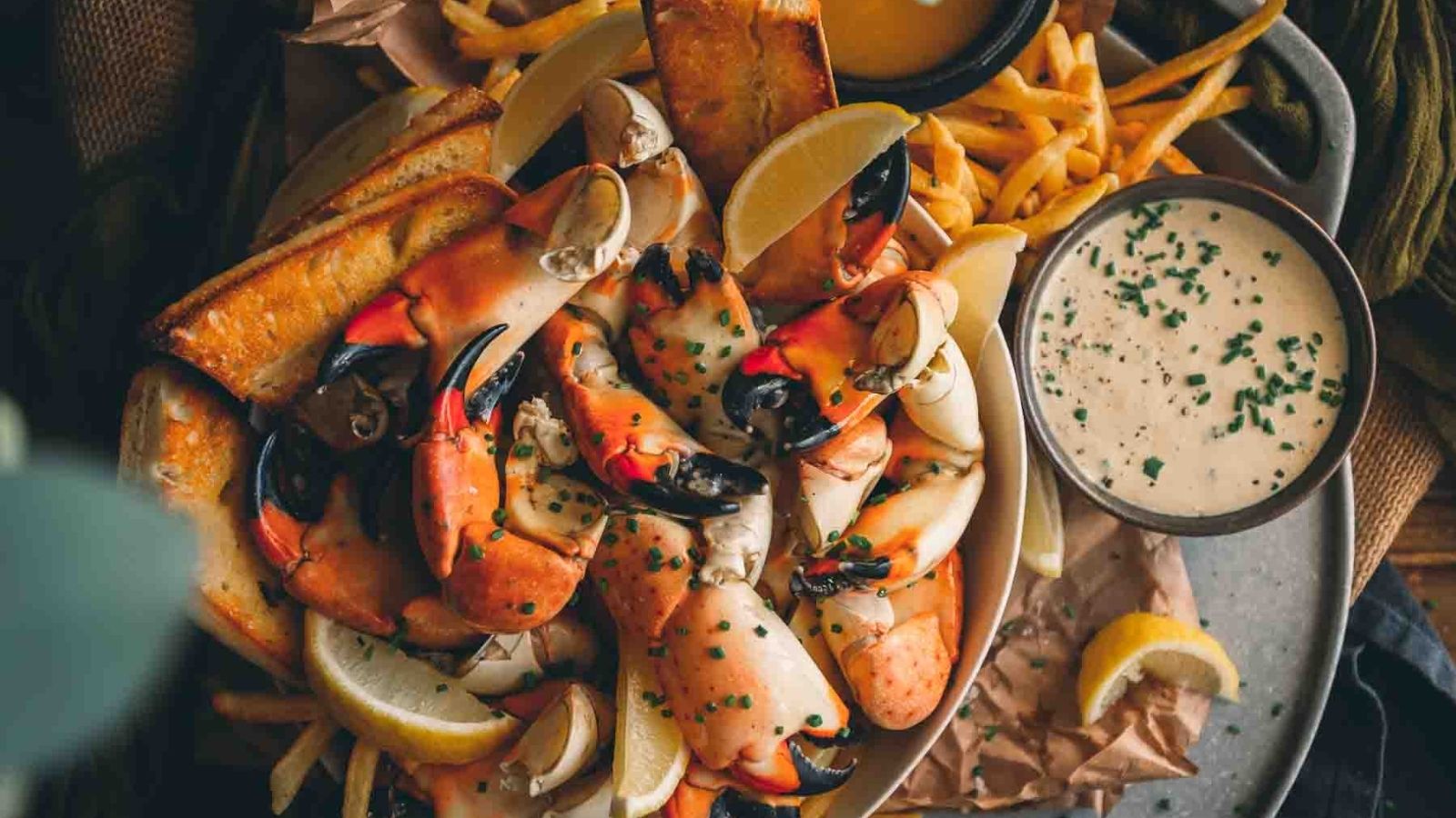 A platter of seafood claws with lemon wedges, crispy fries, toasted bread, and a small bowl of creamy dipping sauce.