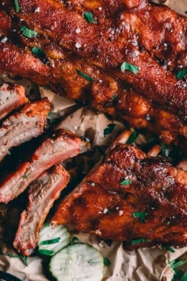 Barbecued ribs on parchment paper with sliced pieces and some herbs sprinkled on top, accompanied by cucumber slices.