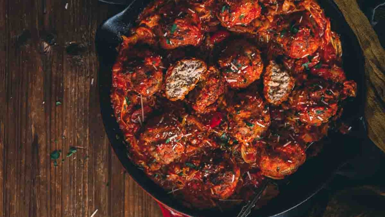 A cast iron skillet filled with meatballs in a rich tomato sauce, garnished with herbs and grated cheese, sits on a wooden surface.