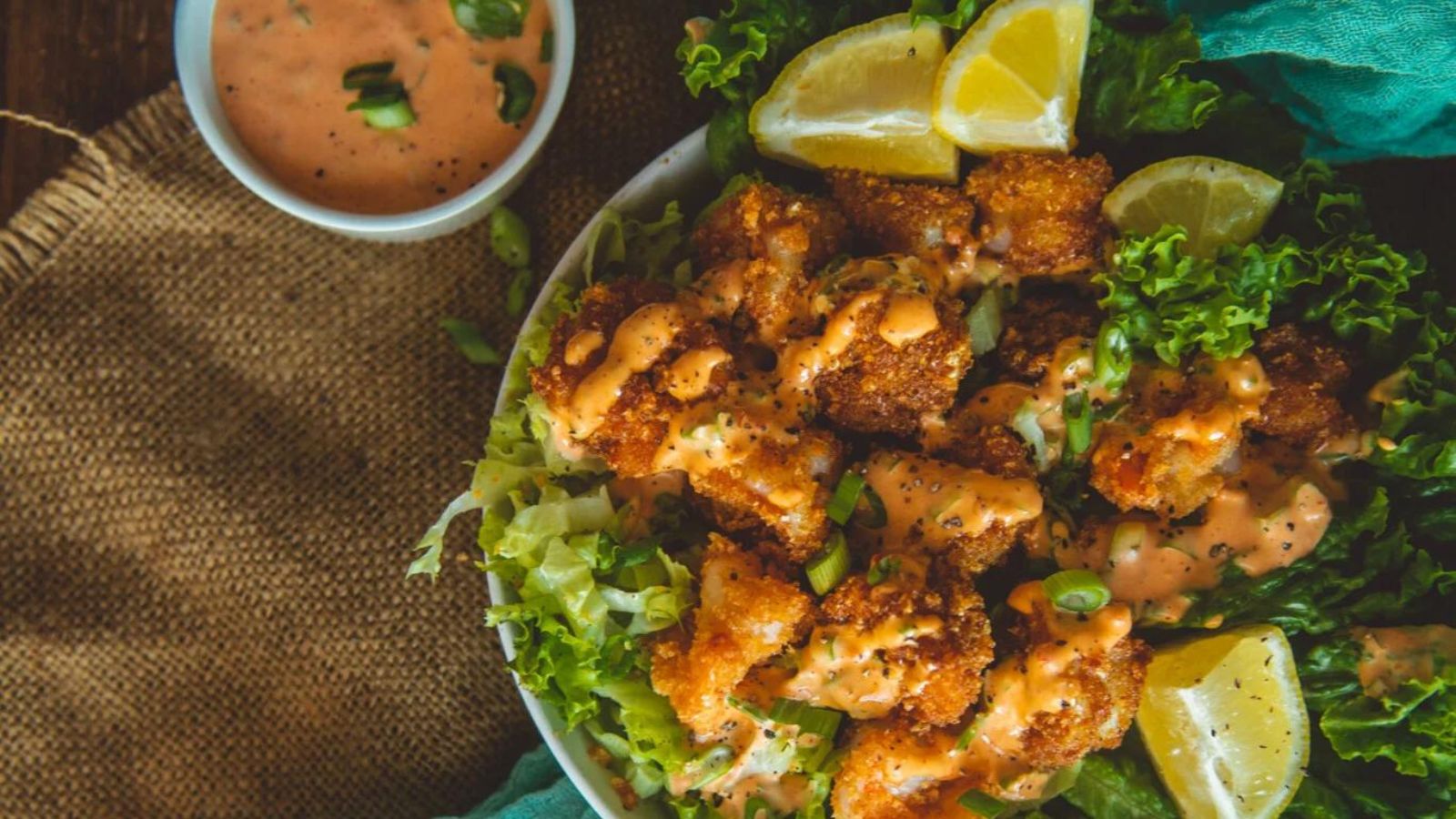 A plate of fried chicken pieces on a bed of lettuce topped with sauce, garnished with lemon wedges. A small bowl of similar sauce is on the side.