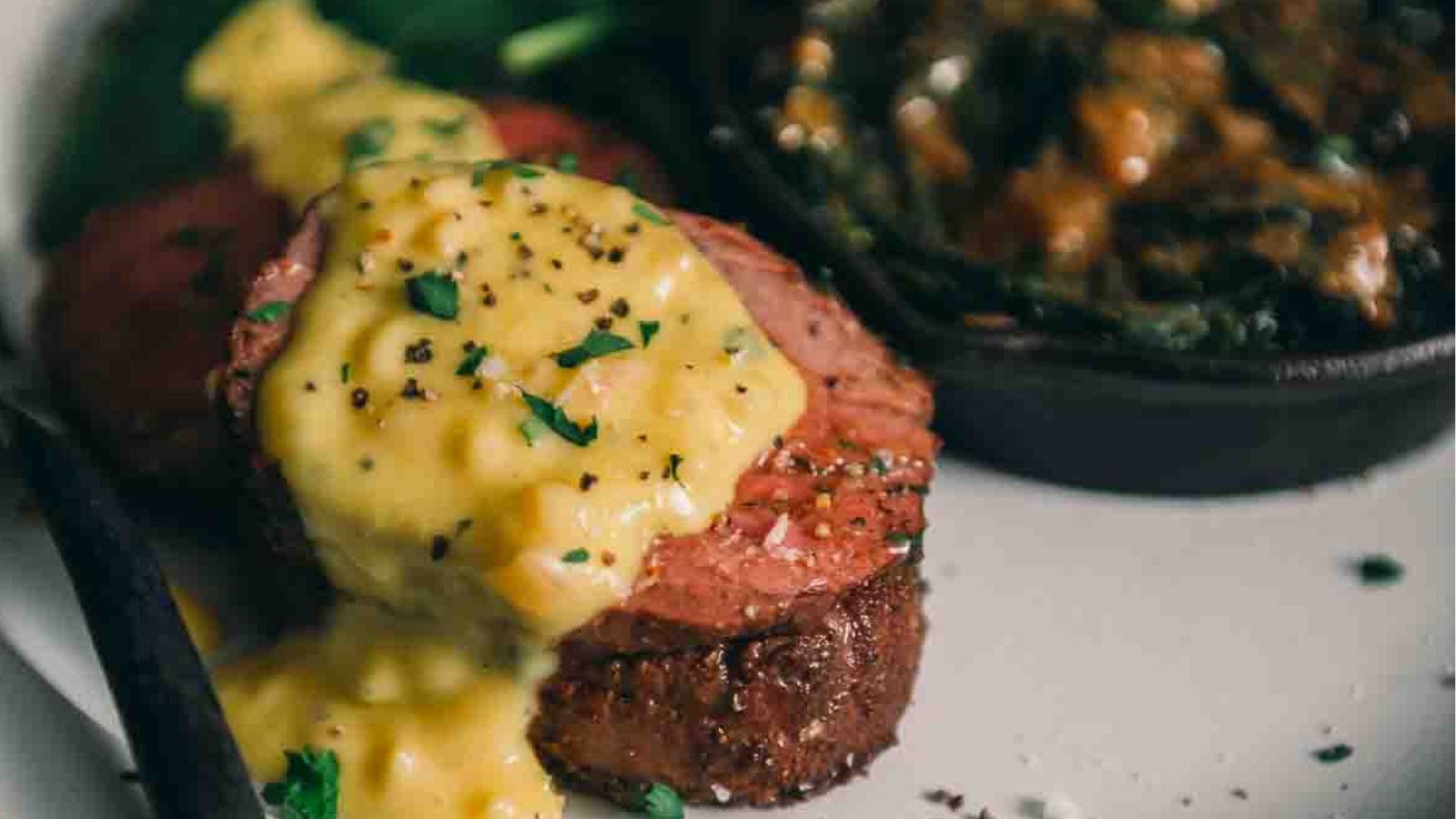 A plate with sliced beef tenderloin topped with one of the best creamy sauces and herbs, served alongside a dish of greens.