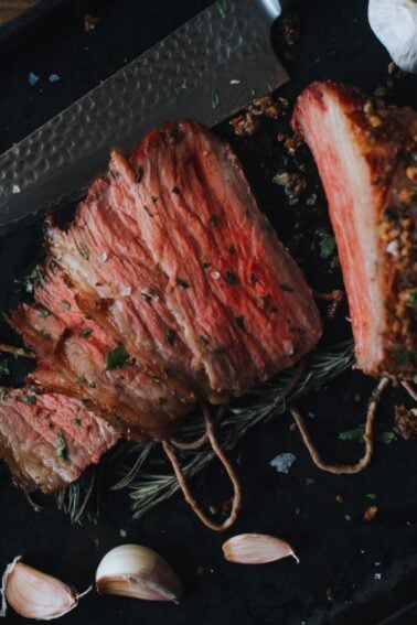 Sliced cooked beef on a black tray with garlic cloves and a knife nearby.