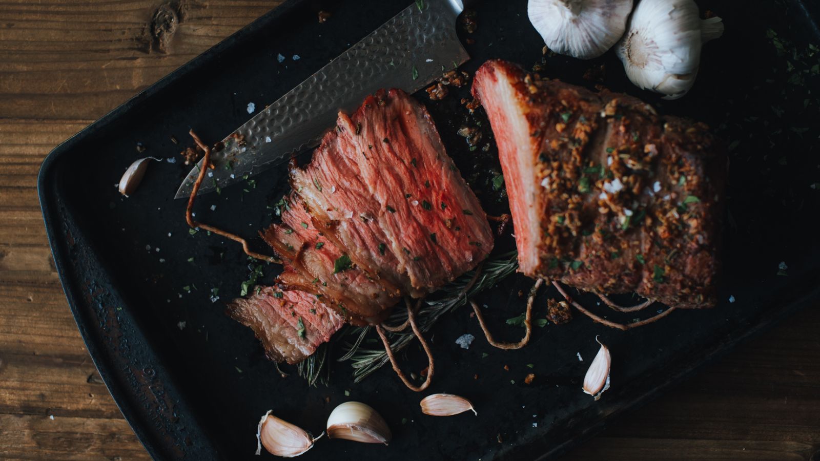 Sliced cooked beef on a black tray with garlic cloves and a knife nearby.