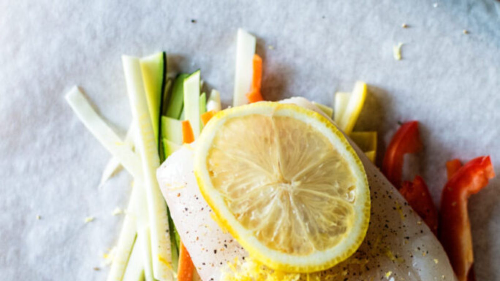 Slice of fish with a lemon on top, surrounded by sliced zucchini, carrots, and red bell peppers on parchment paper.