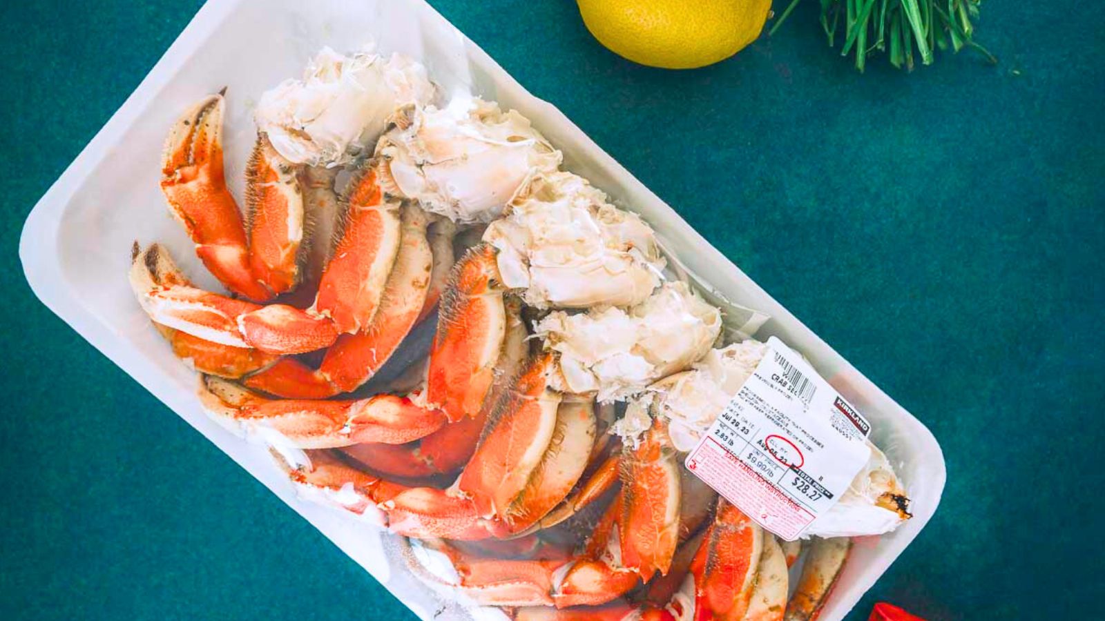 Packaged cooked crab legs in a plastic container on a green surface, surrounded by a lemon and herbs.