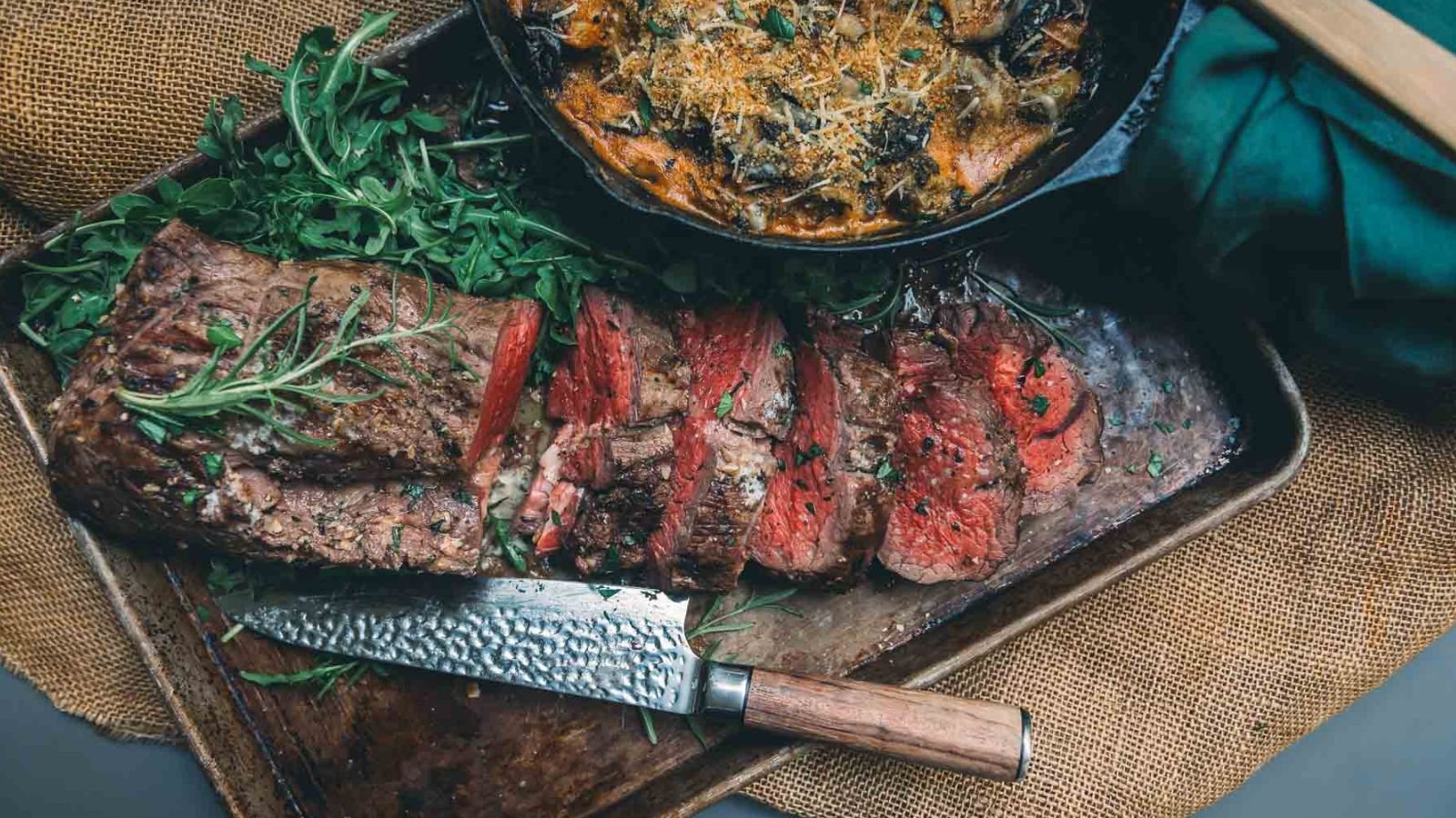 Sliced roasted beef on a tray with a sprig of rosemary, beside a cast iron pan of cooked vegetables, garnished with herbs. A knife with a wooden handle rests nearby, ready to cut the perfect portion to pair with the best sauces for beef tenderloin.