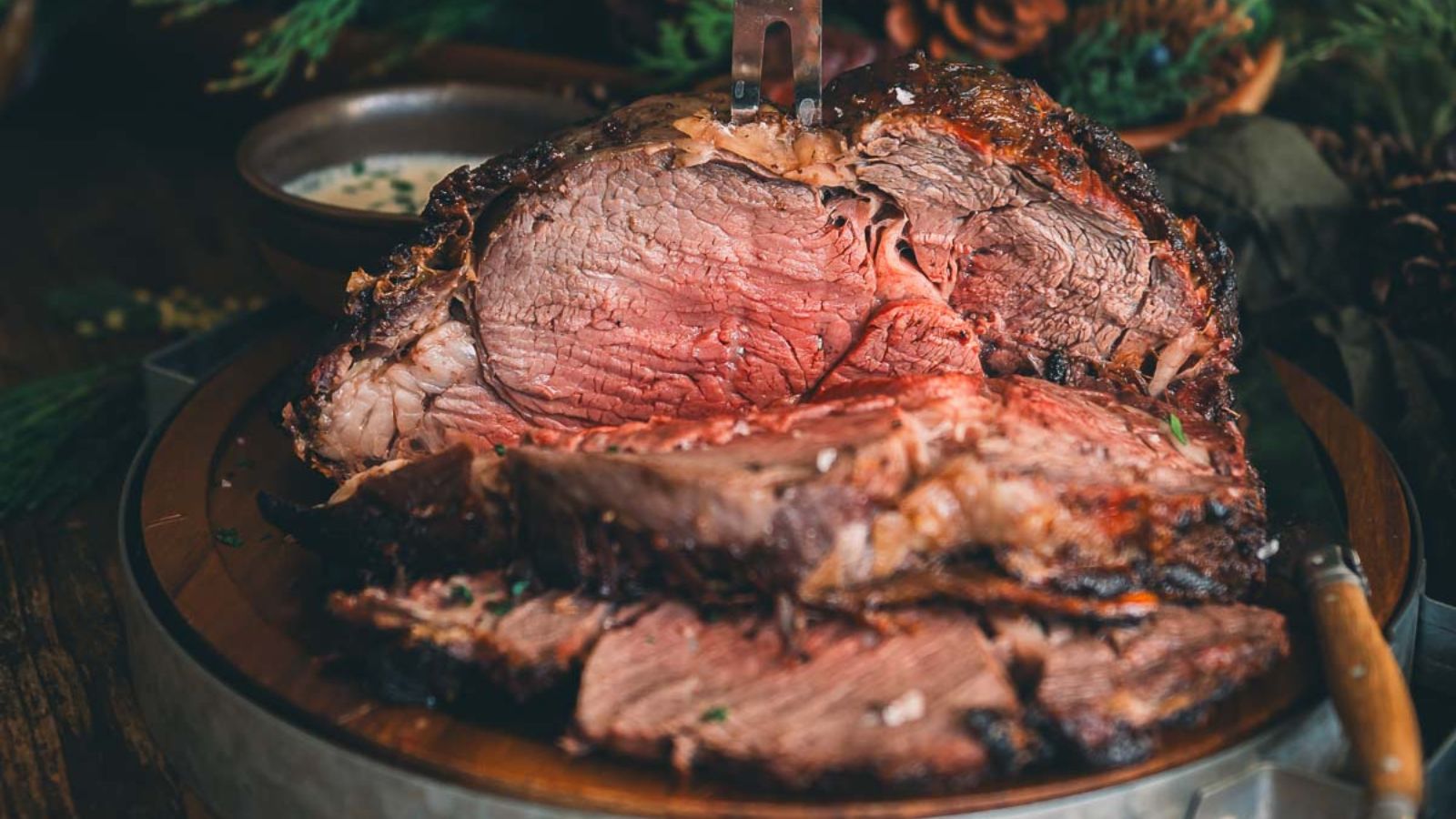 A large, roasted beef cut with a pink, juicy center is displayed on a wooden platter, accompanied by a side of sauce in a small bowl.