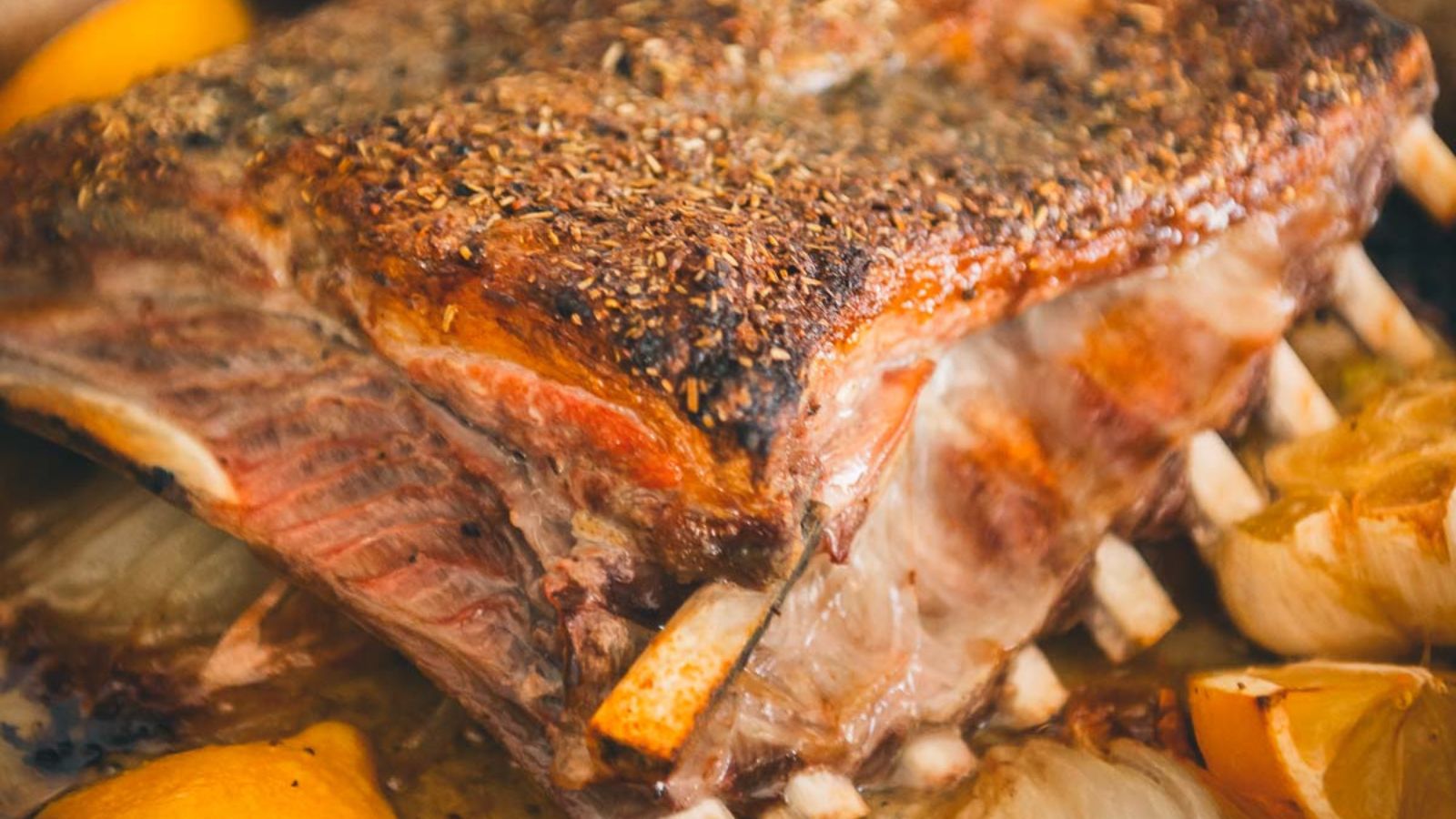 Close-up of a seasoned, roasted rack of ribs with visible herbs and spices, surrounded by lemon wedges and onions.