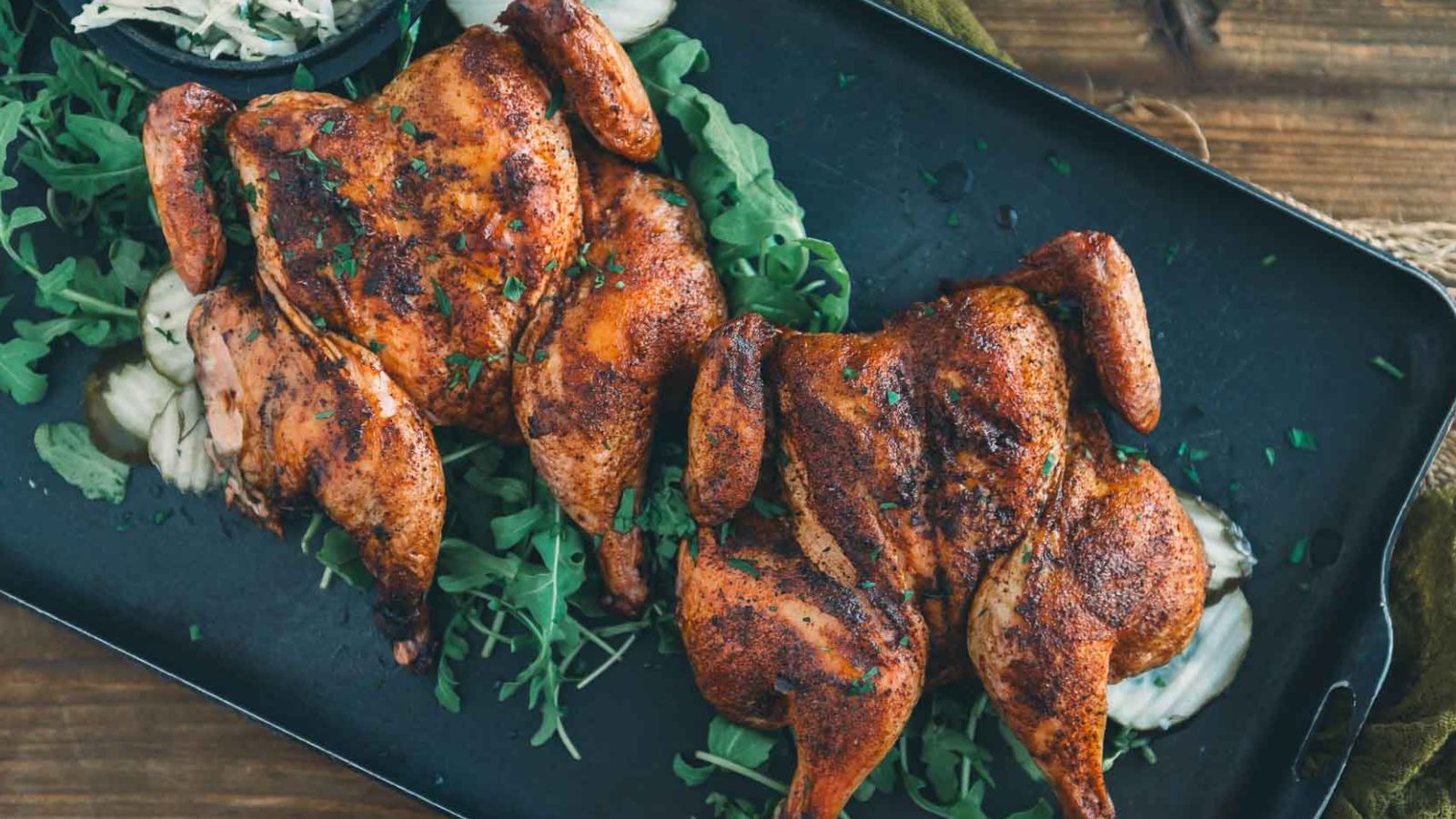 Two roasted spatchcock chickens on a tray, garnished with fresh greens and sliced onions.