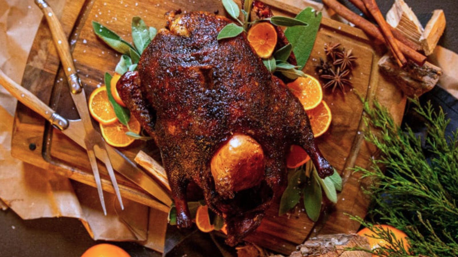 Roasted duck garnished with orange slices, herbs, and spices on a wooden cutting board. Silver carving tools and rustic decor are visible nearby.