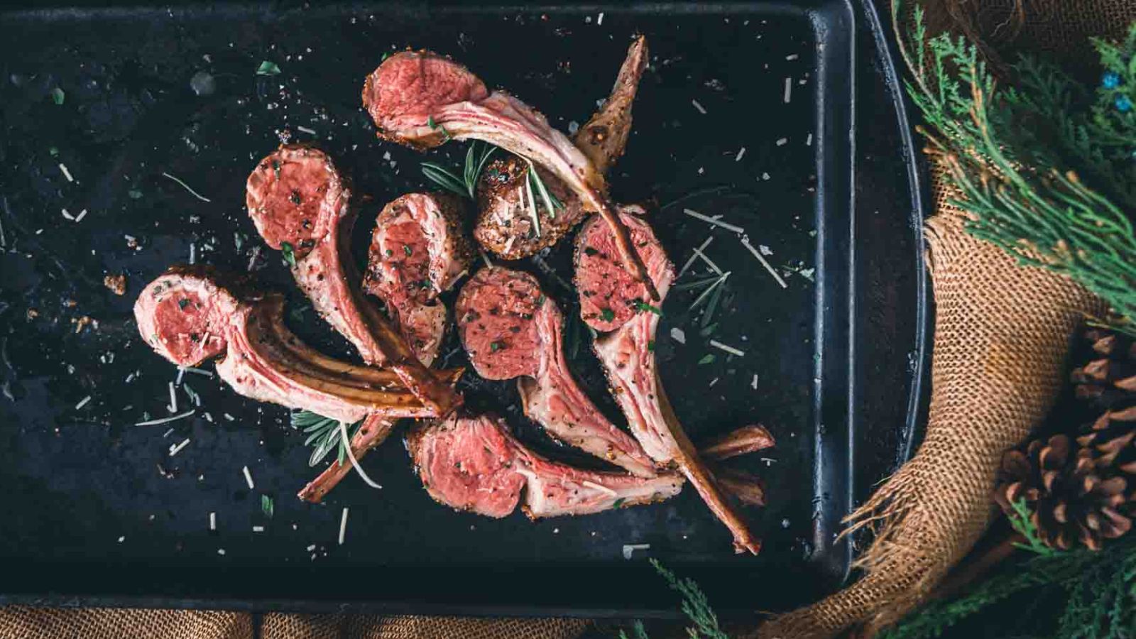 Grilled lamb chops seasoned with herbs on a dark tray, placed on a rustic fabric with greenery around.