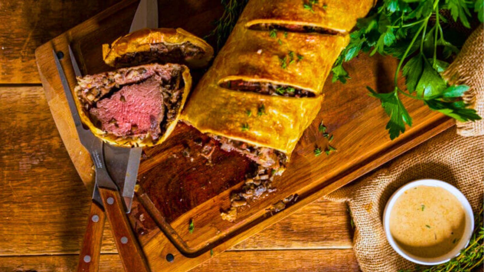 A sliced beef Wellington on a wooden board with a steak knife, garnished with herbs. Beside it sits a small bowl of the best sauce for beef tenderloin, enhancing every bite.