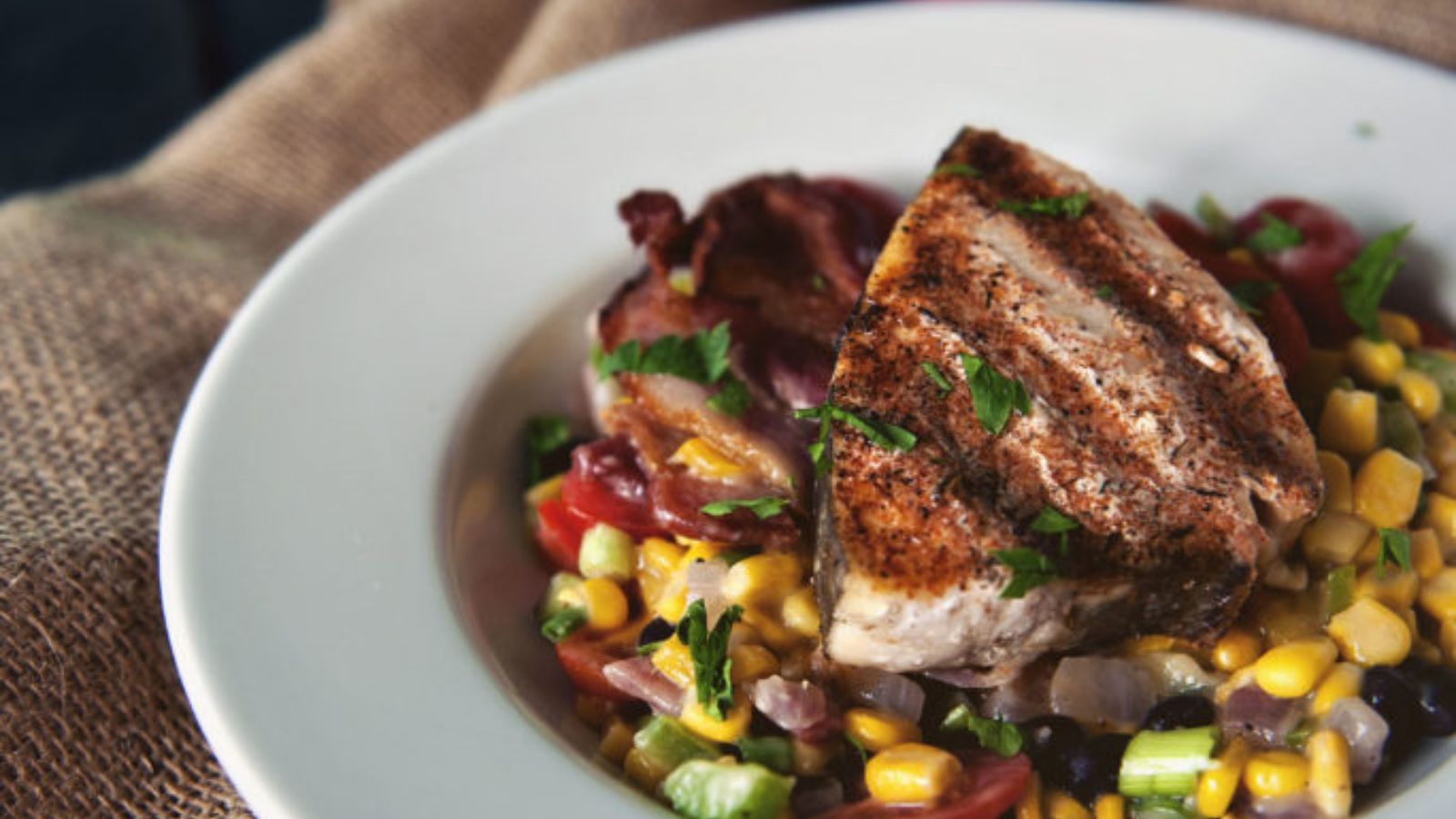 Grilled fish fillet on top of a bed of corn, black beans, red onions, tomatoes, and herbs, served on a white plate.