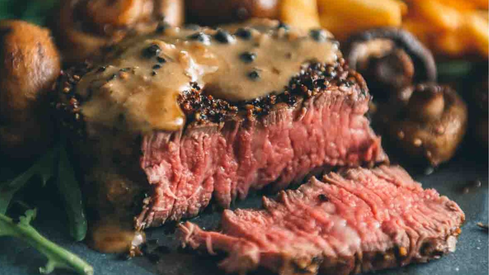 Close-up of a sliced medium-rare steak topped with one of the best sauces for beef tenderloin, a rich peppercorn blend, accompanied by mushrooms and leafy greens.