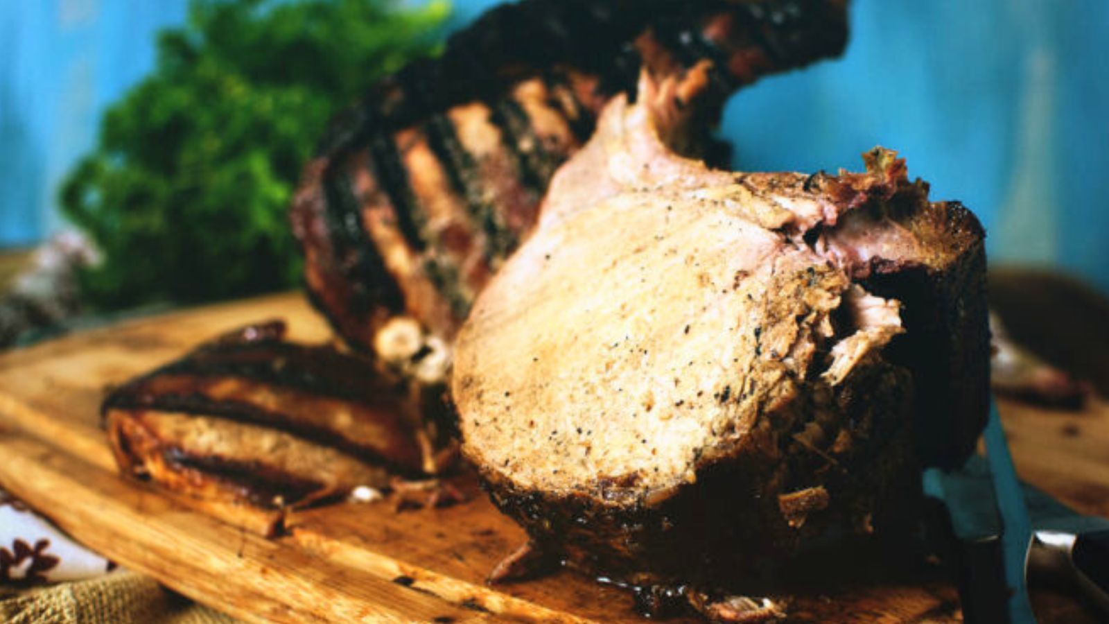 A sliced roast pork with visible grill marks on a wooden cutting board, accompanied by greens in the background.