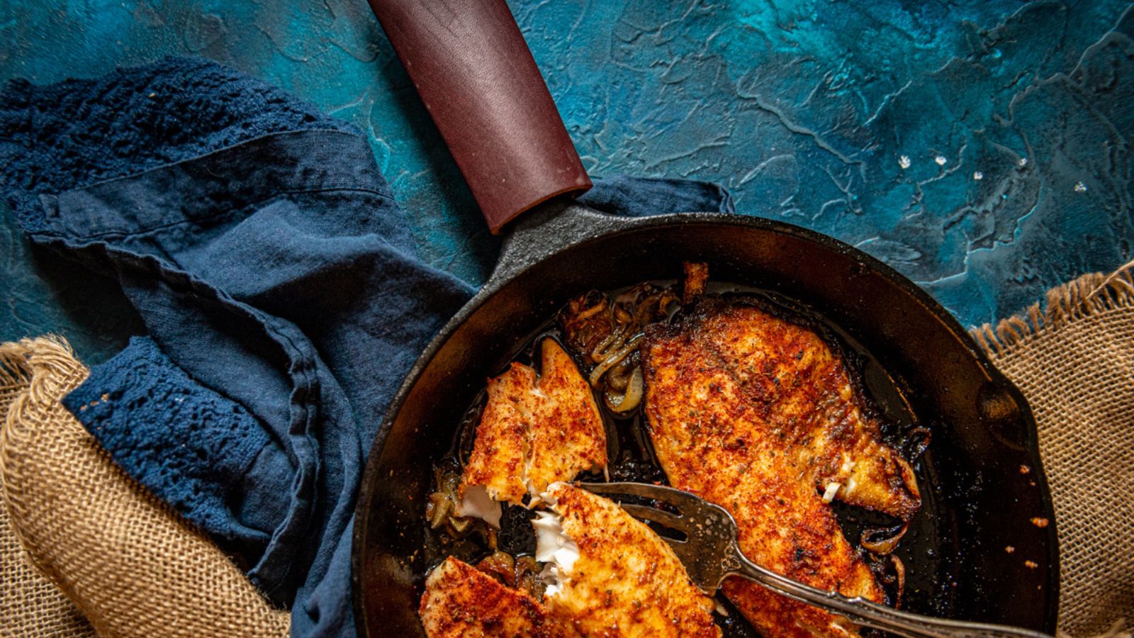 A skillet with cooked, seasoned fish fillets on a blue surface, accompanied by a dark blue cloth and burlap.