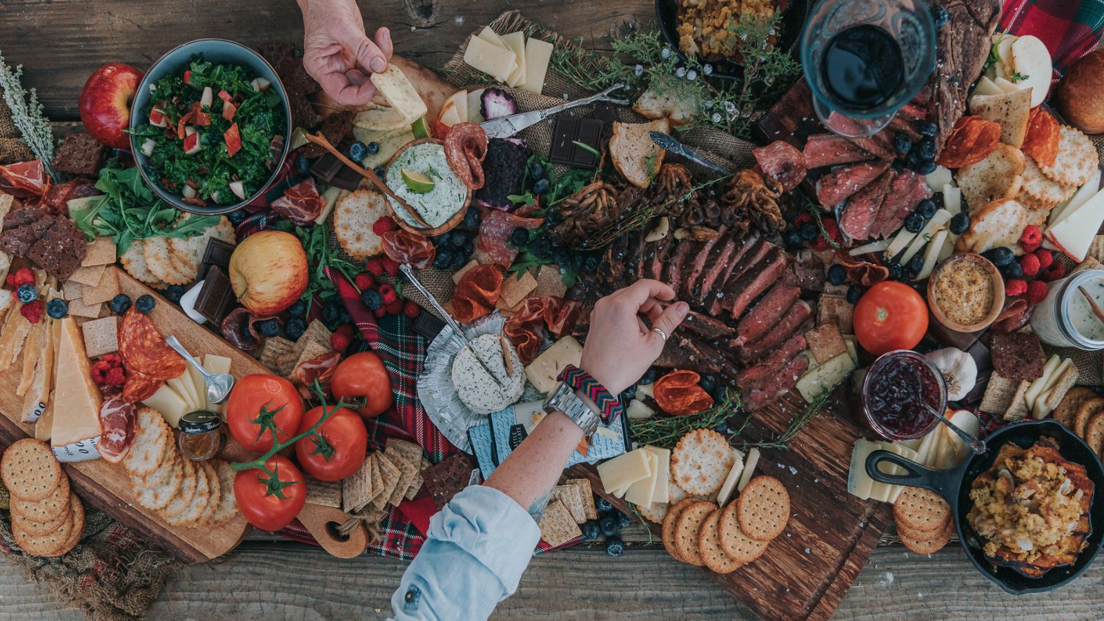 A variety of foods on a table, including cheeses, meats, fruits, tomatoes, bread, and salad. Hands are reaching for items, and a person pours wine.