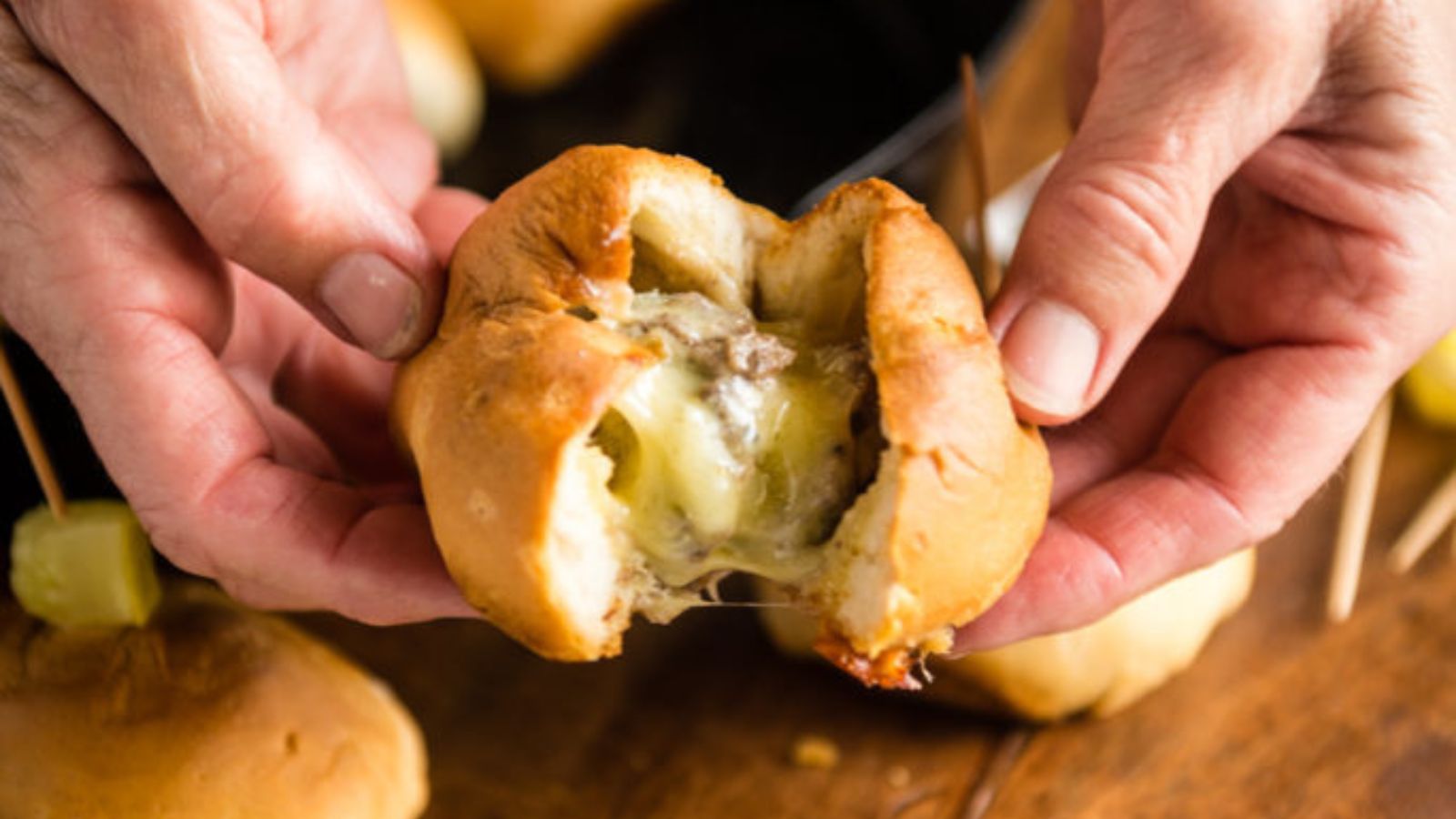 Hands holding a cooked bread roll filled with melted cheese and meat.