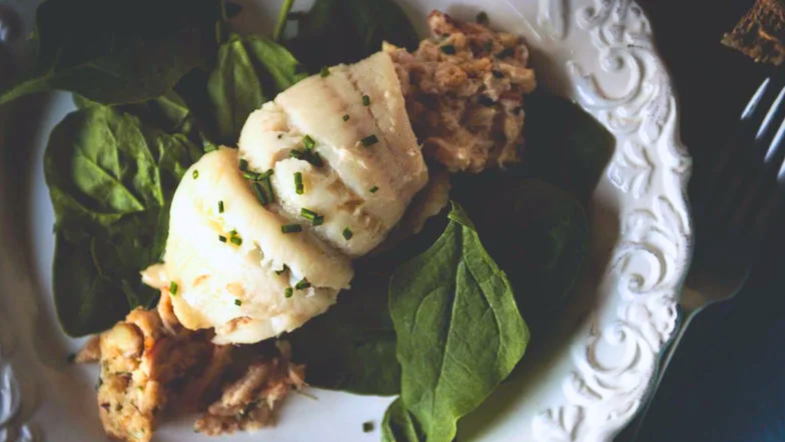 A plate with rolled white fish on a bed of spinach, garnished with herbs, and a side of filling, on a decorative white plate.