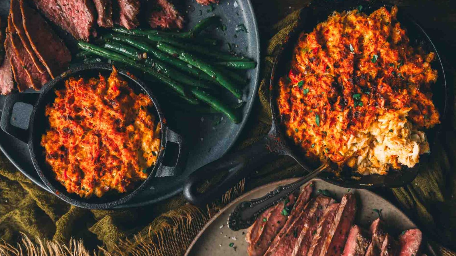 Plates of sliced steak, green beans, and baked dishes topped with melted cheese are displayed on a table.