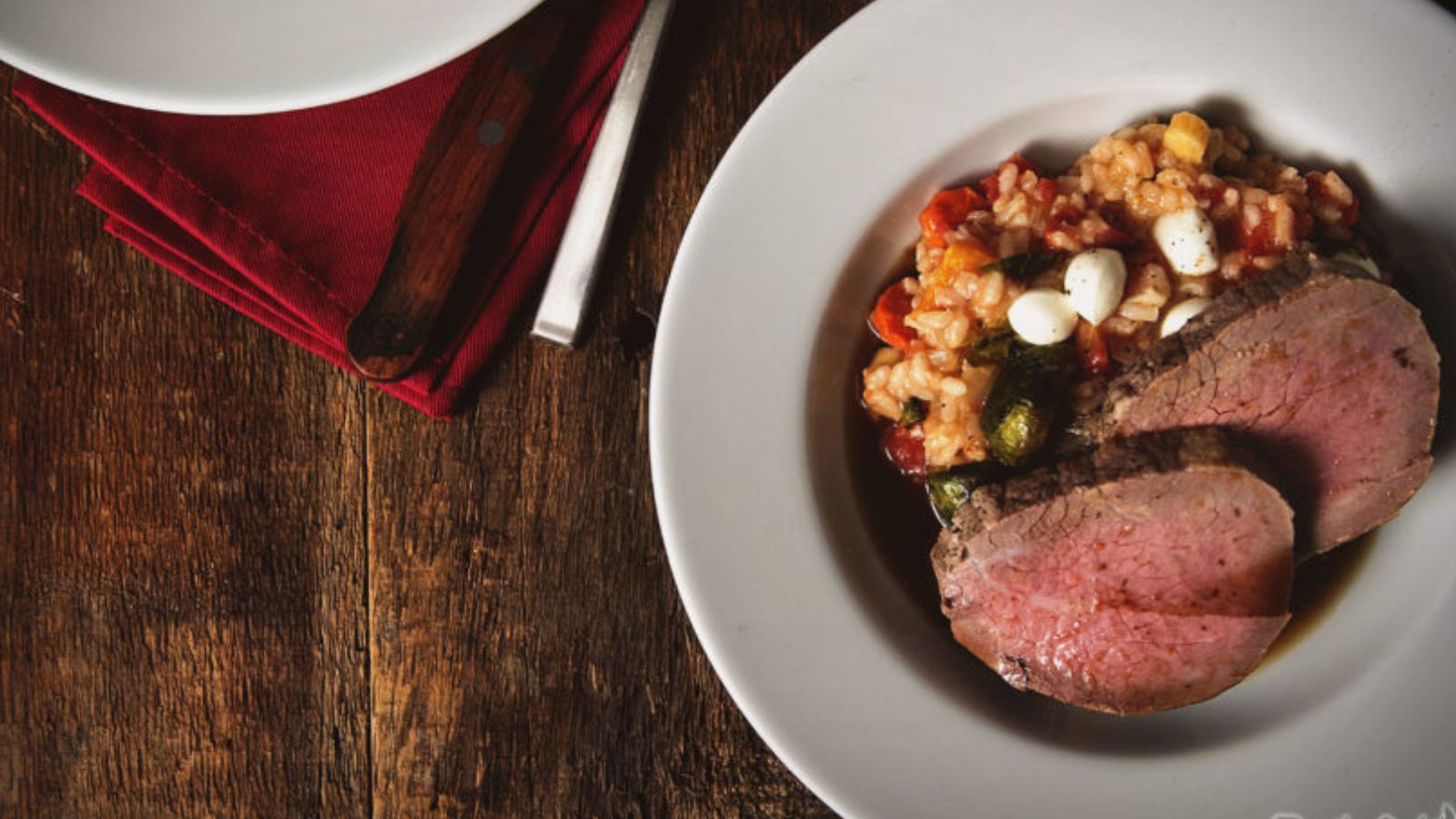 A plate of sliced roast beef with vegetables and rice on a wooden table. Red napkin and fork on the side.