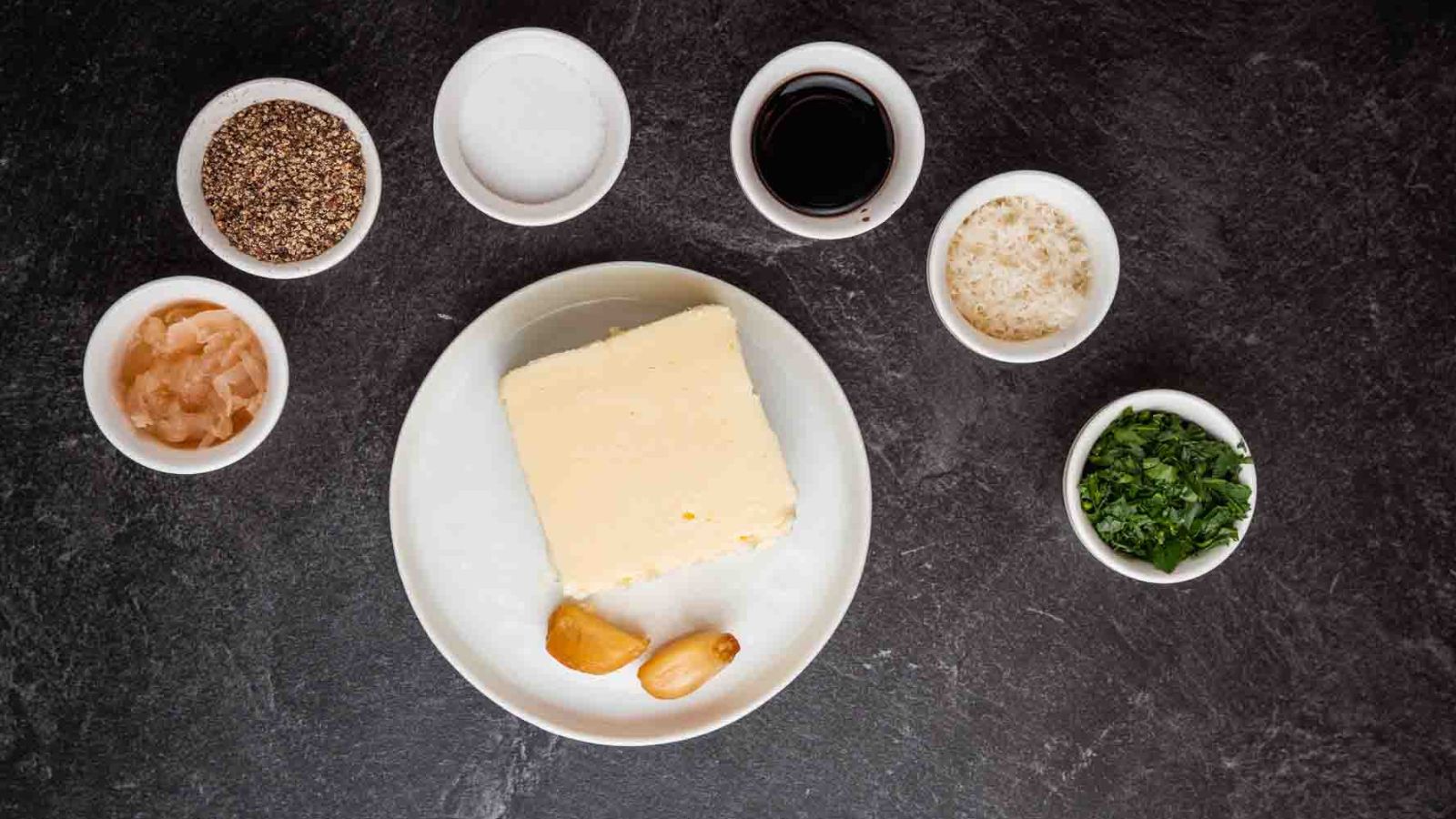 A block of butter and two garlic cloves rest on a plate, surrounded by small bowls of spices and herbs—ideal ingredients for crafting the best sauces for beef tenderloin—all elegantly arranged on a dark surface.