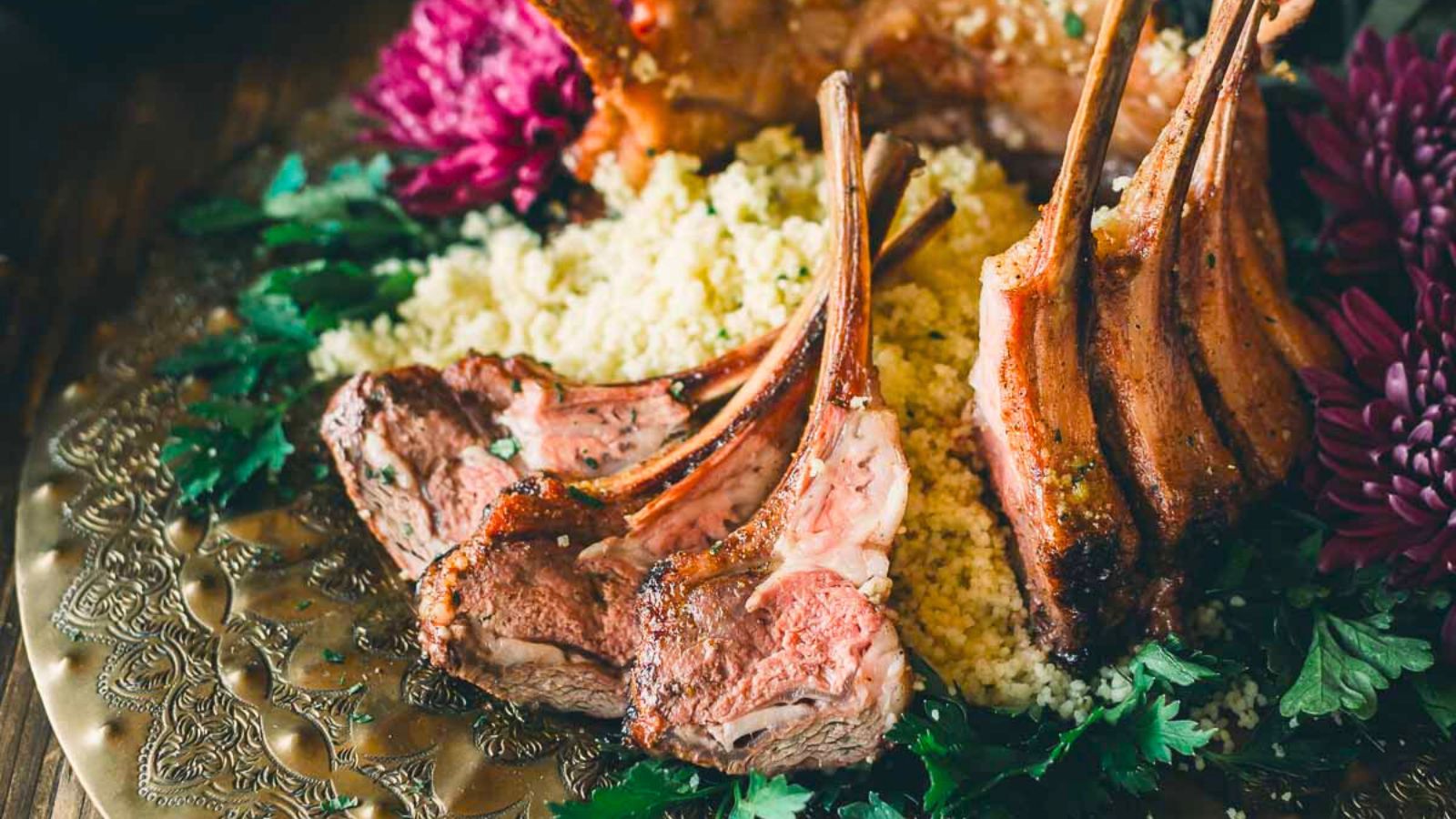 Roasted rack of lamb on a bed of couscous, garnished with fresh herbs and purple flowers on an ornate plate.