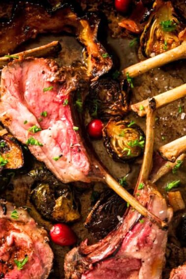 Grilled rack of lamb with some slices cut, garnished with herbs, surrounded by roasted vegetables and cranberries.