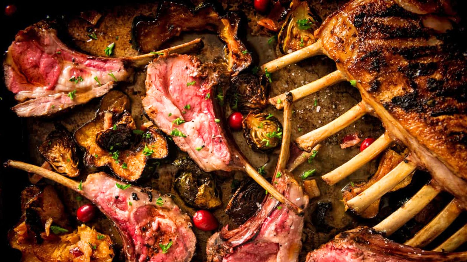 Grilled rack of lamb with some slices cut, garnished with herbs, surrounded by roasted vegetables and cranberries.