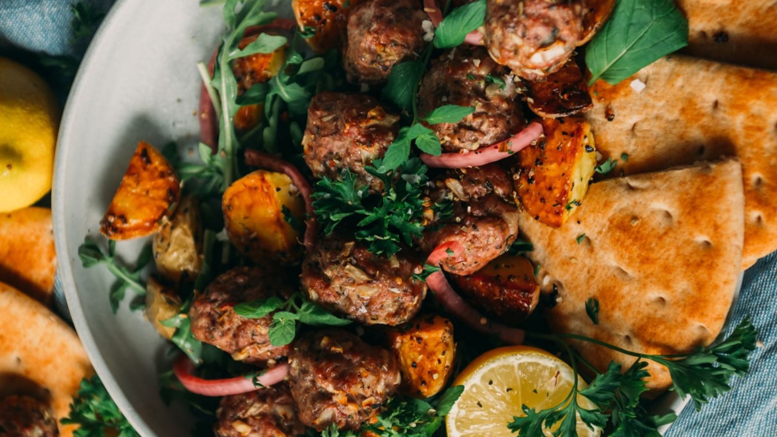 A plate with grilled meatballs, roasted vegetables, flatbread, and lemon slices garnished with fresh herbs.