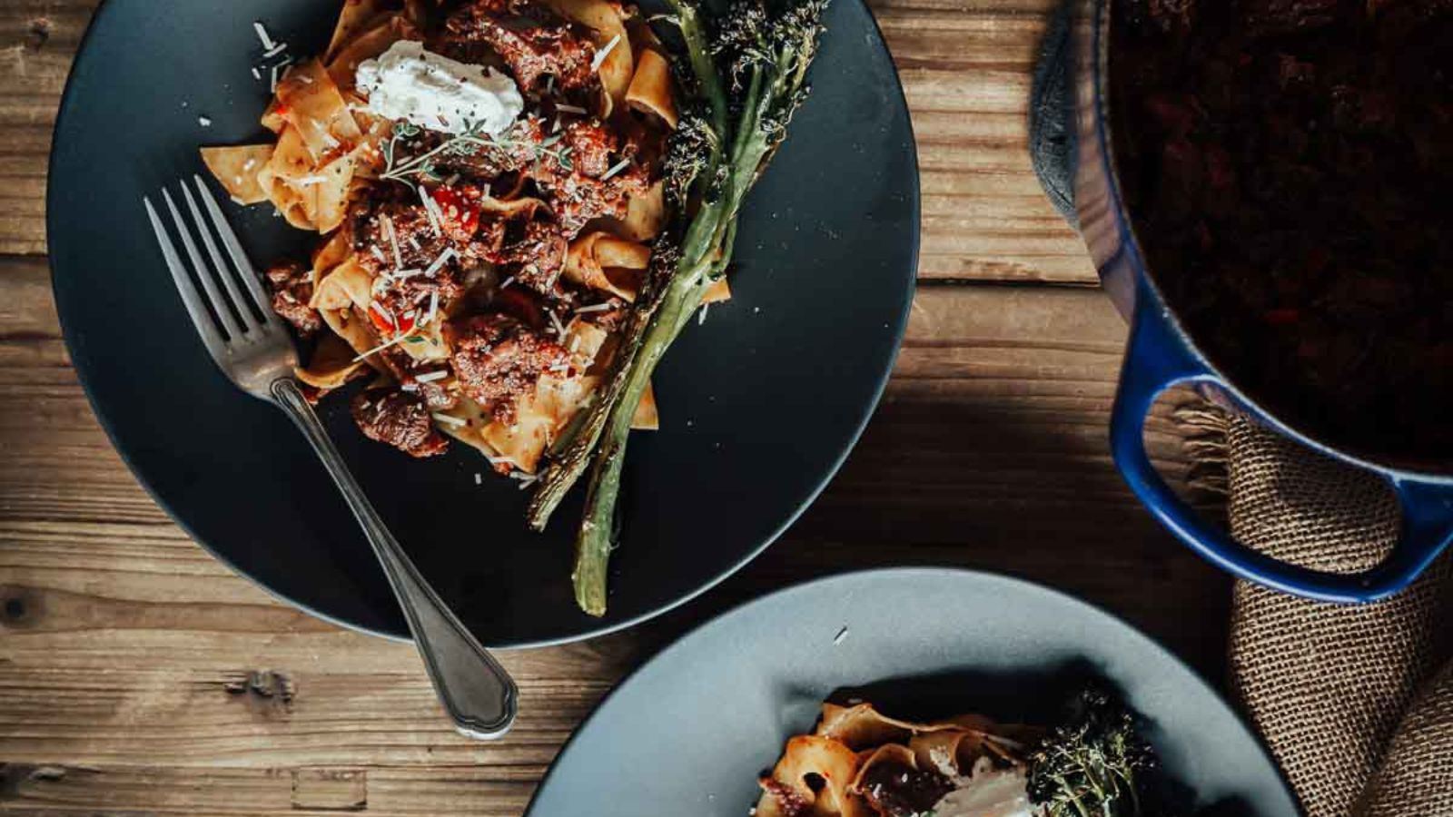 Two plates of pasta with meat sauce, garnished with asparagus and herbs, sit on a wooden table next to a blue pot.