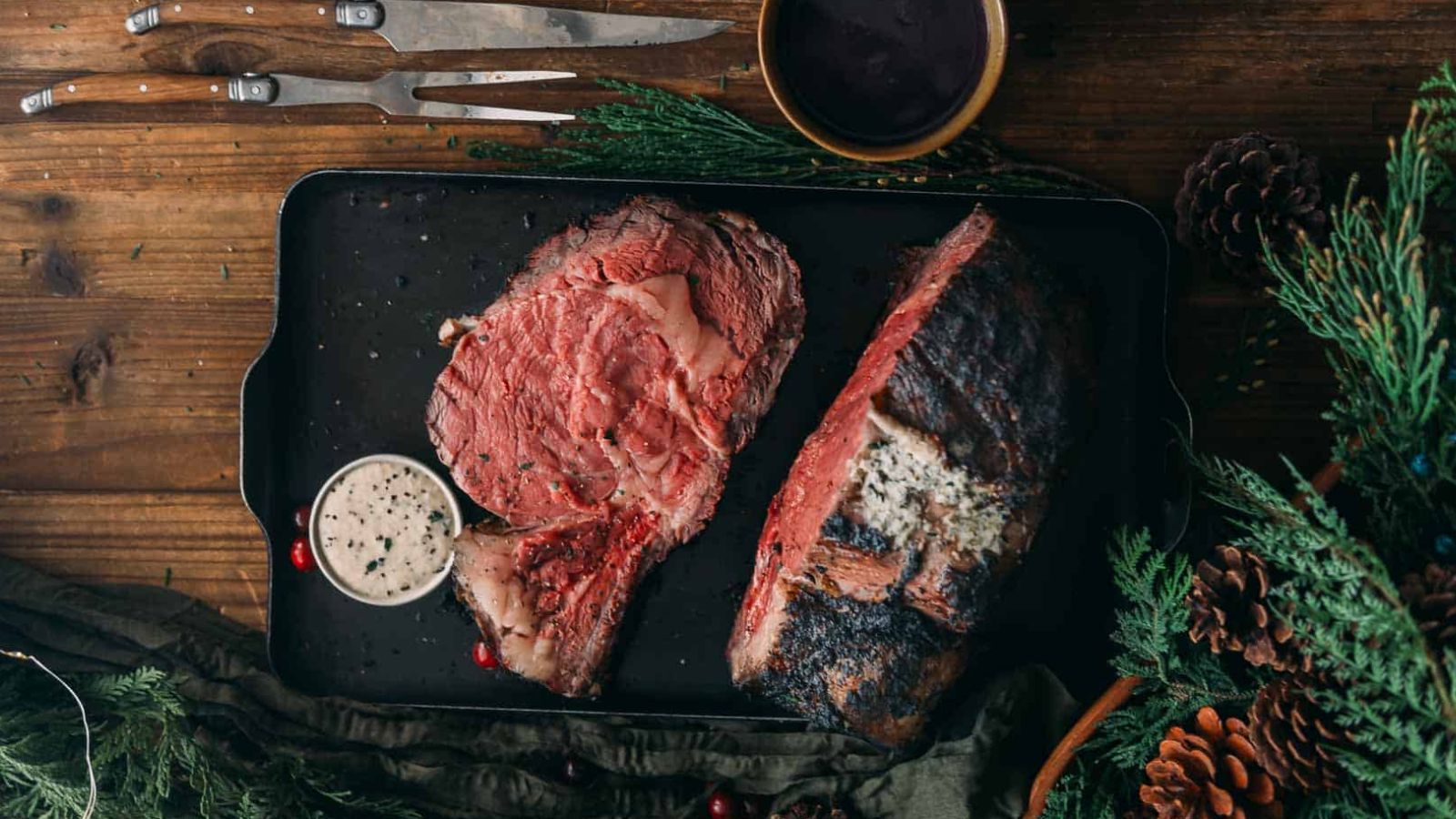 A tray with cooked prime rib, two meat forks, a bowl of sauce, and evergreen sprigs on a wooden table.