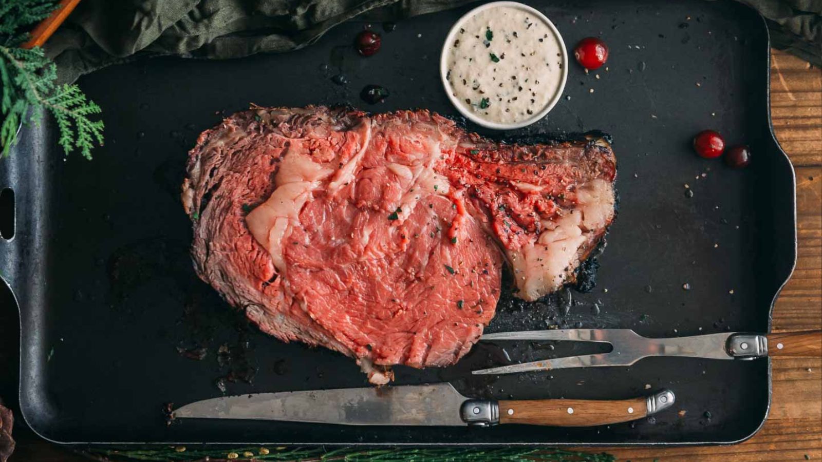 Slice of medium-rare prime rib on a black tray with a bowl of sauce, garnished with cranberries and herbs.