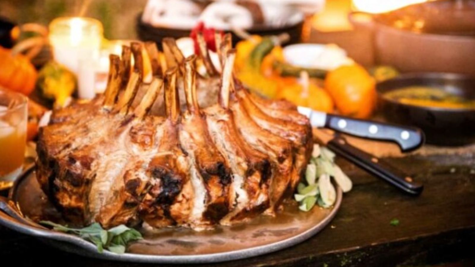 A cooked rack of lamb is placed on a platter garnished with herbs. The background includes lit candles and small pumpkins.