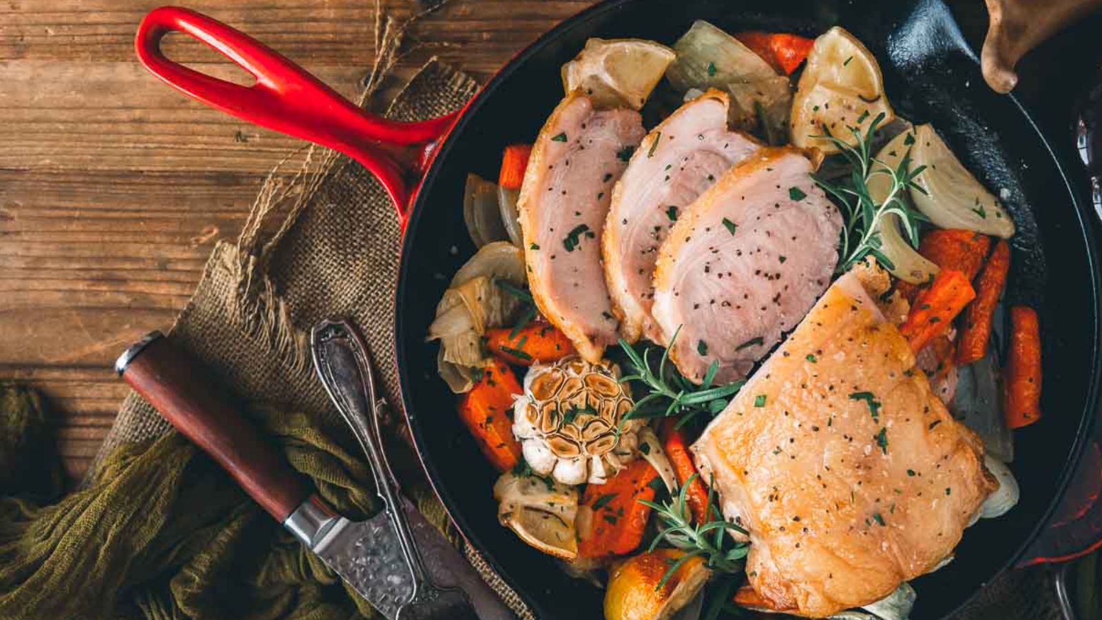 A skillet with slices of cooked meat, carrots, onions, and lemon wedges, garnished with fresh rosemary.