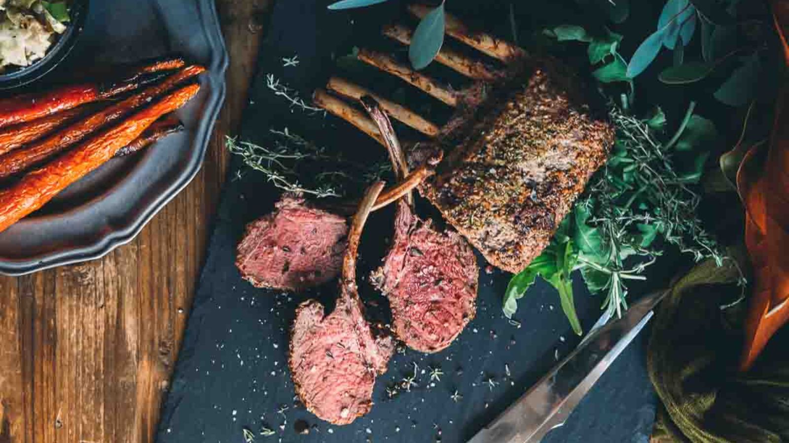 Sliced rack of lamb on a slate board, garnished with herbs, with cooked carrots on the side.