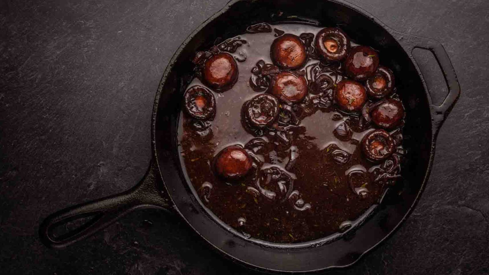 A cast iron pan sizzles with mushrooms and onions immersed in one of the best sauces for beef tenderloin, creating a rich aroma on a sleek black surface.