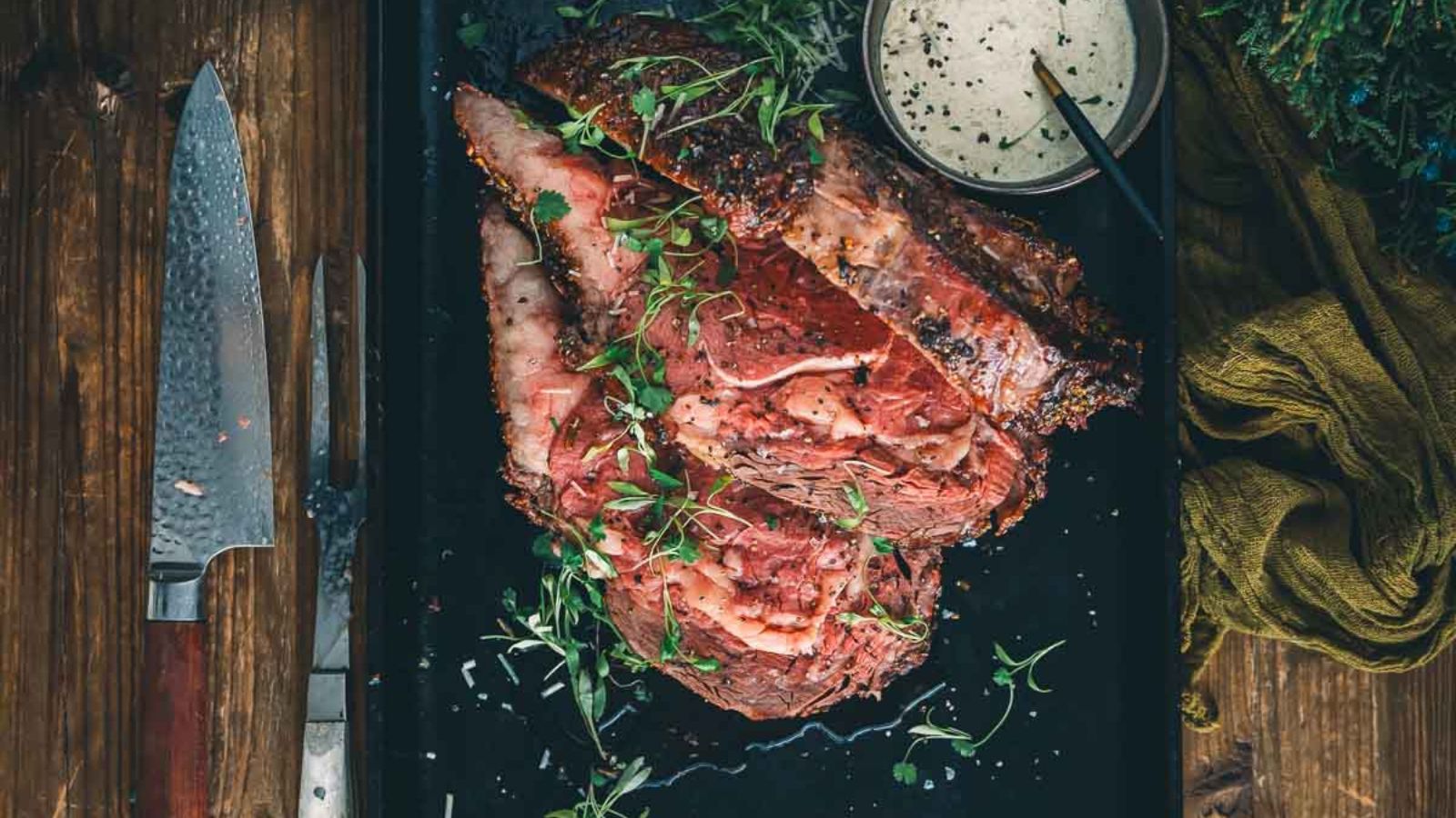 A tray with sliced roast beef garnished with herbs, a knife on the side, and a bowl of creamy sauce.