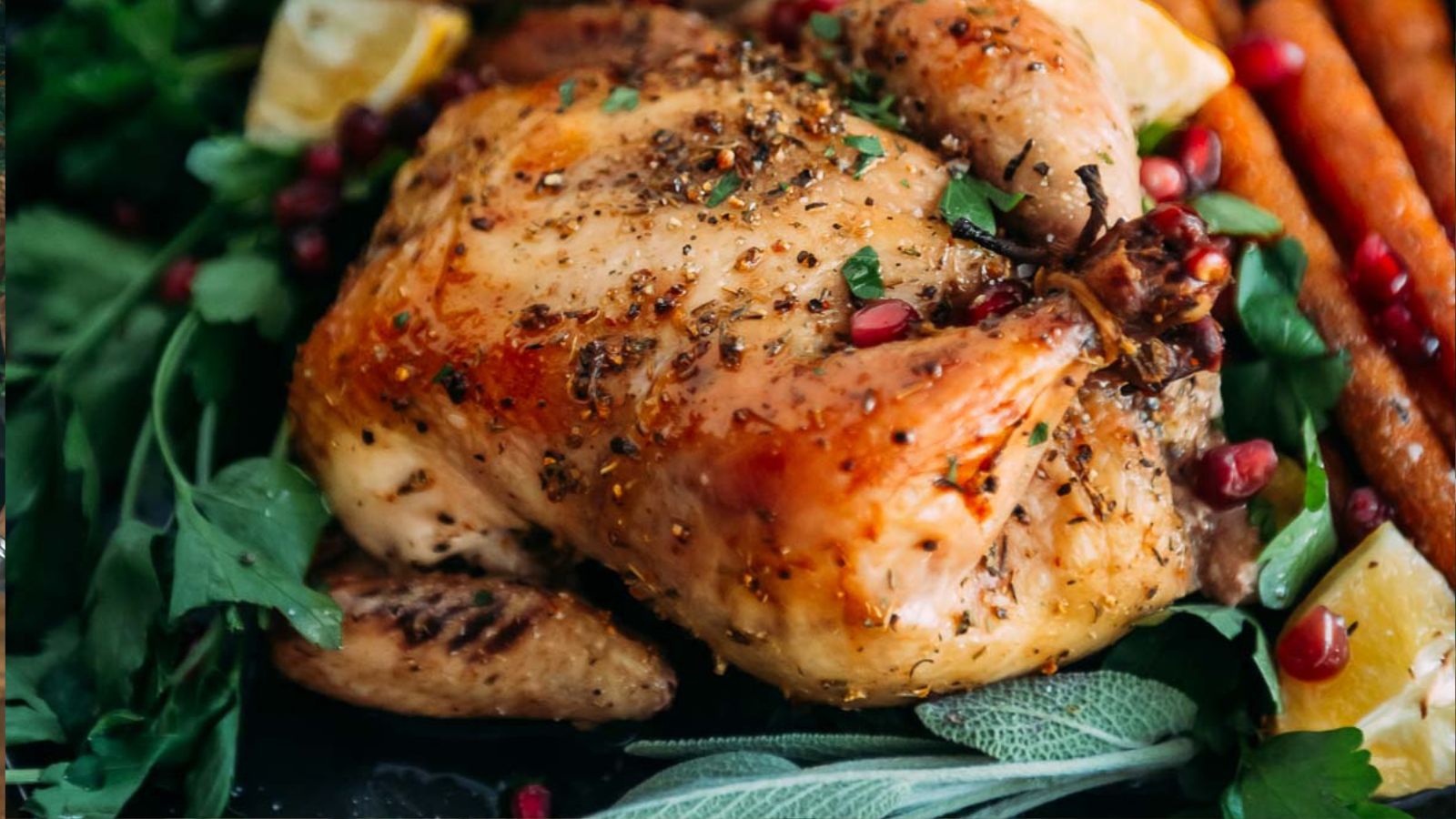 Roasted chicken garnished with herbs, lemon wedges, pomegranate seeds, and surrounded by carrots.