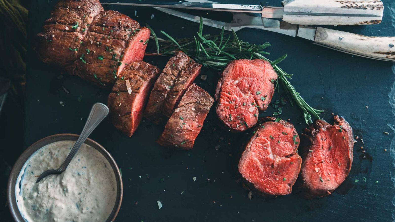 Sliced medium-rare roast beef on a dark surface, garnished with herbs, sits enticingly next to a bowl of one of the best sauces for beef tenderloin and a set of knives.