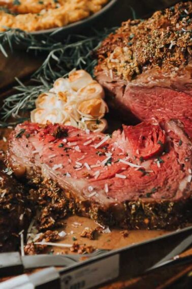 Sliced roasted beef on a tray, surrounded by herbs and a whole head of roasted garlic, with a bowl of sauce in the background.