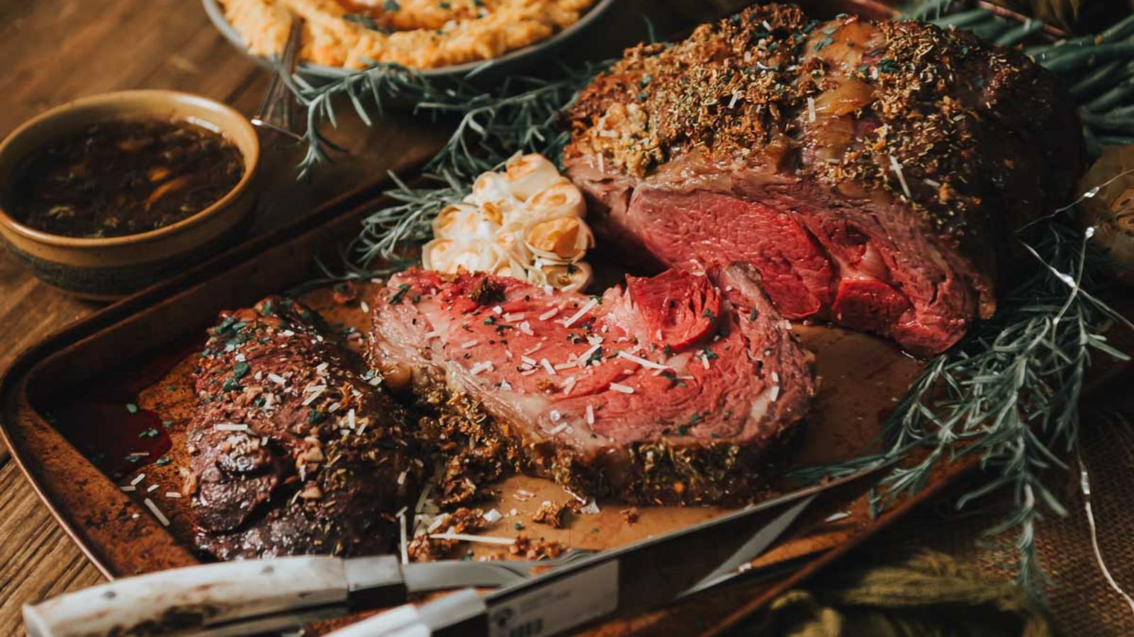 Sliced roasted beef on a tray, surrounded by herbs and a whole head of roasted garlic, with a bowl of sauce in the background.