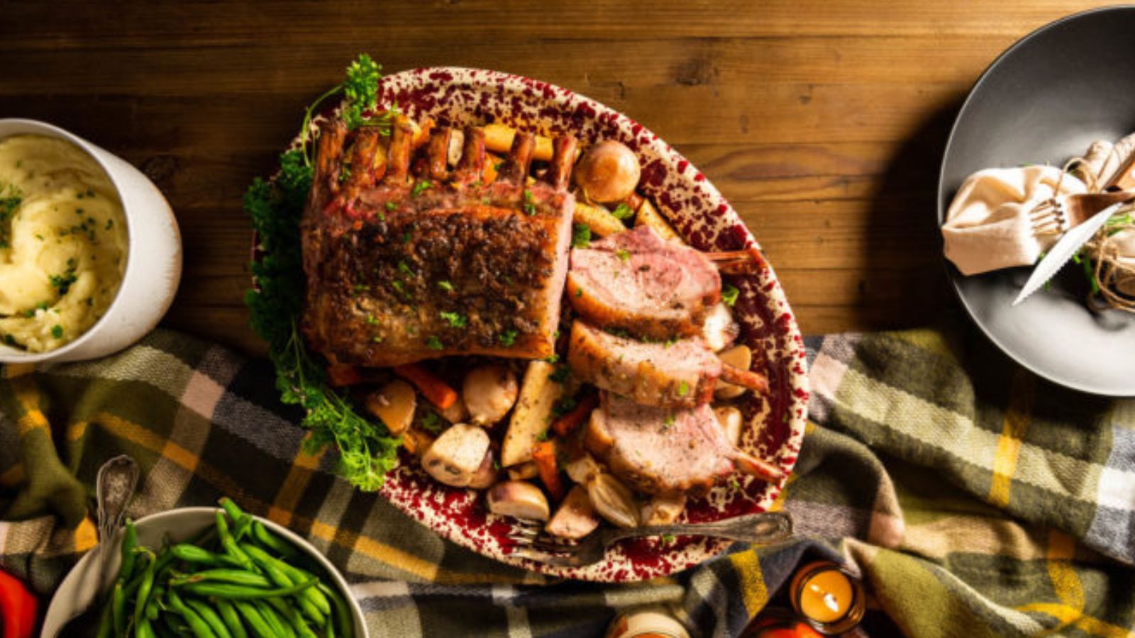 A platter of roast pork with herbs, surrounded by potatoes and mushrooms, is on a wooden table with mashed potatoes, green beans, and a napkin on the side.