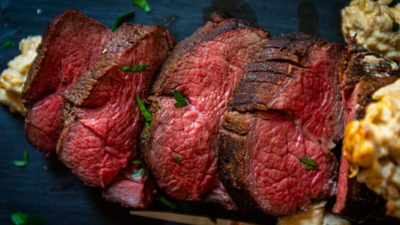 Slices of medium-rare beef tenderloin rest on a dark surface, garnished with parsley and ready for the best sauces.