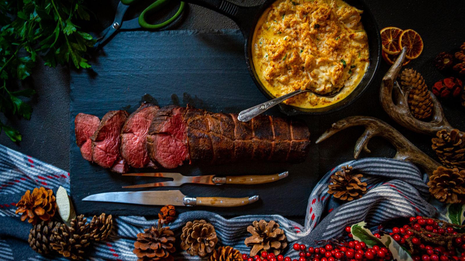 Sliced roast beef sits next to gratin in a skillet, surrounded by pine cones, berries, and a striped cloth. Discovering the best sauces for beef tenderloin could elevate this feast even further.