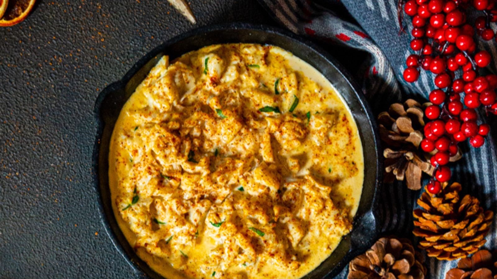 A creamy cauliflower dish, perfect as a Christmas appetizer, is presented in a dark bowl, surrounded by pine cones and red berries on a textured surface.