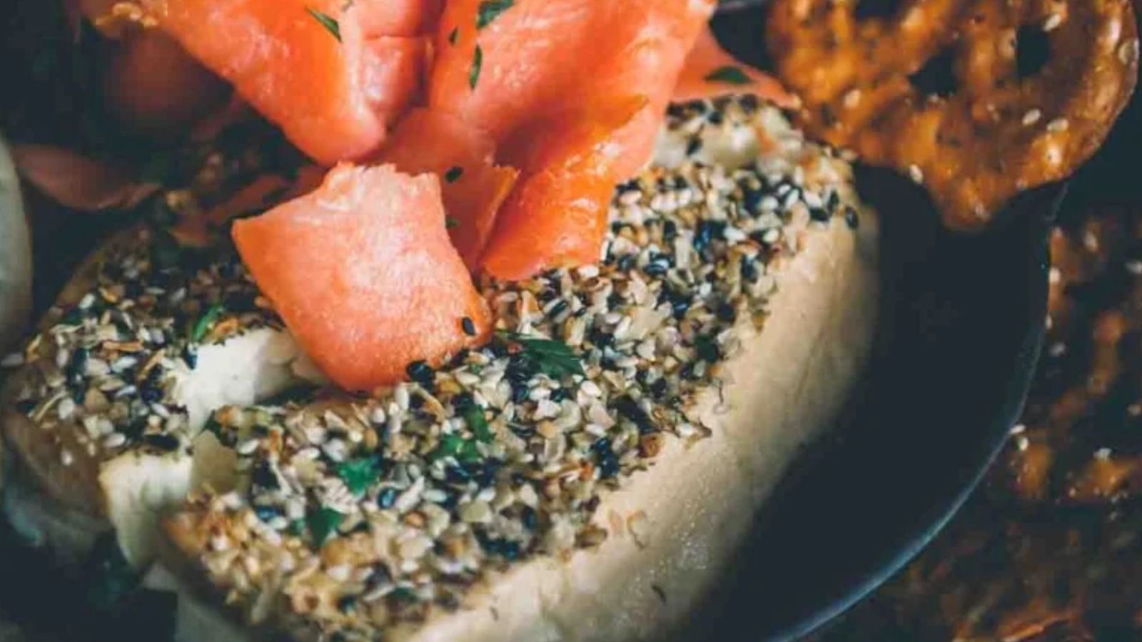 A close-up of a festive bagel topped with herb and seed-coated cream cheese and slices of smoked salmon, perfect as a Christmas appetizer, accompanied by crunchy pretzel snacks.