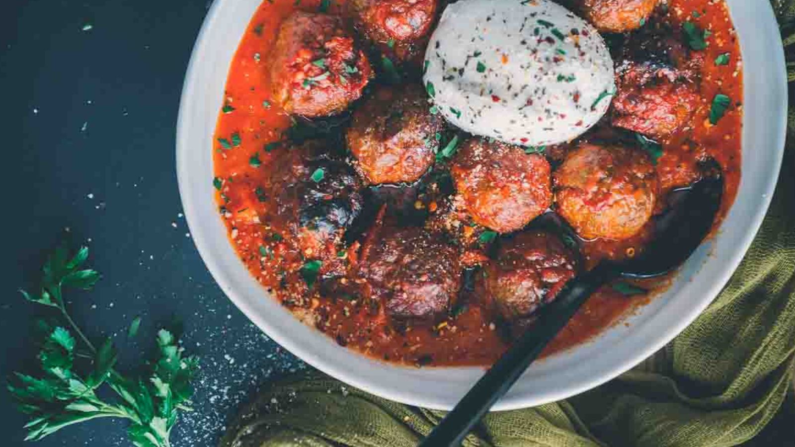 A festive bowl of meatballs in red sauce topped with a dollop of creamy cheese, perfect as Christmas appetizers. Garnished with herbs, a spoon rests invitingly in the bowl.