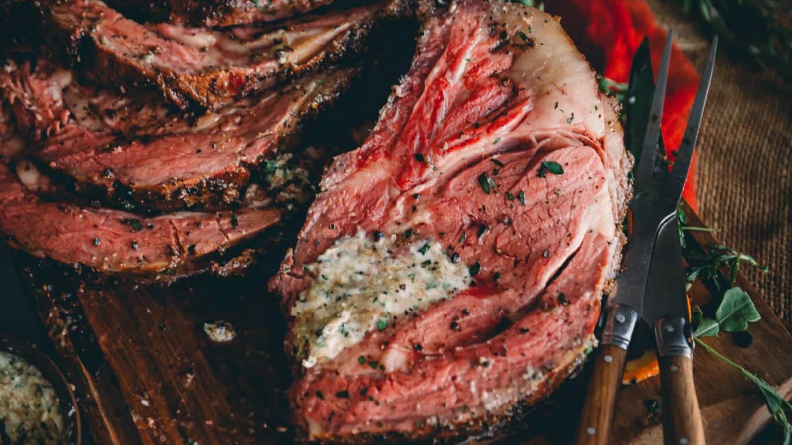 Close-up of a sliced, grilled prime rib with a herb butter coating, garnished with herbs. Fork and knife beside the meat on a wooden board.